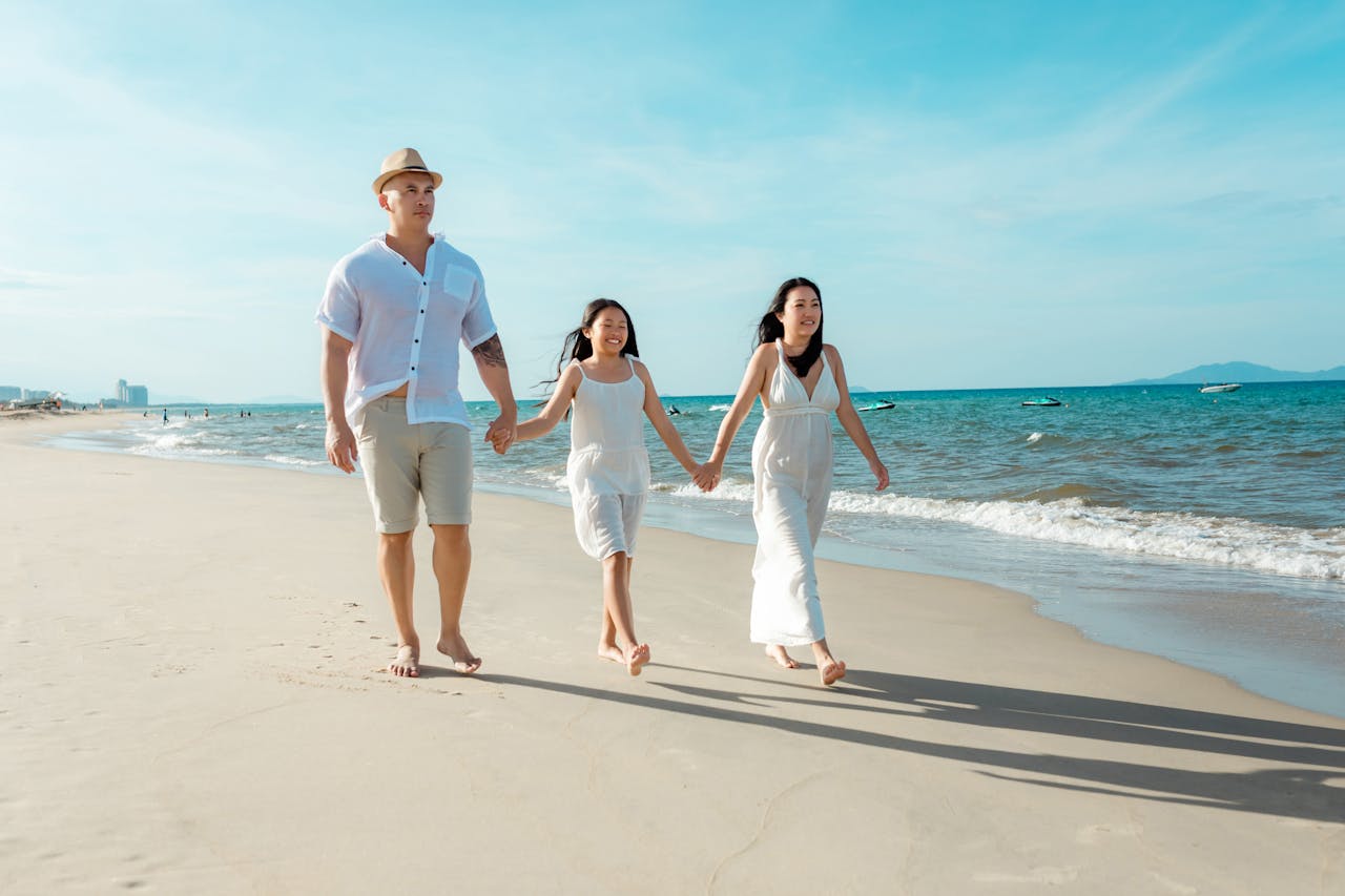 A family strolls along the shore of Hoi An beach on a bright, sunny day.